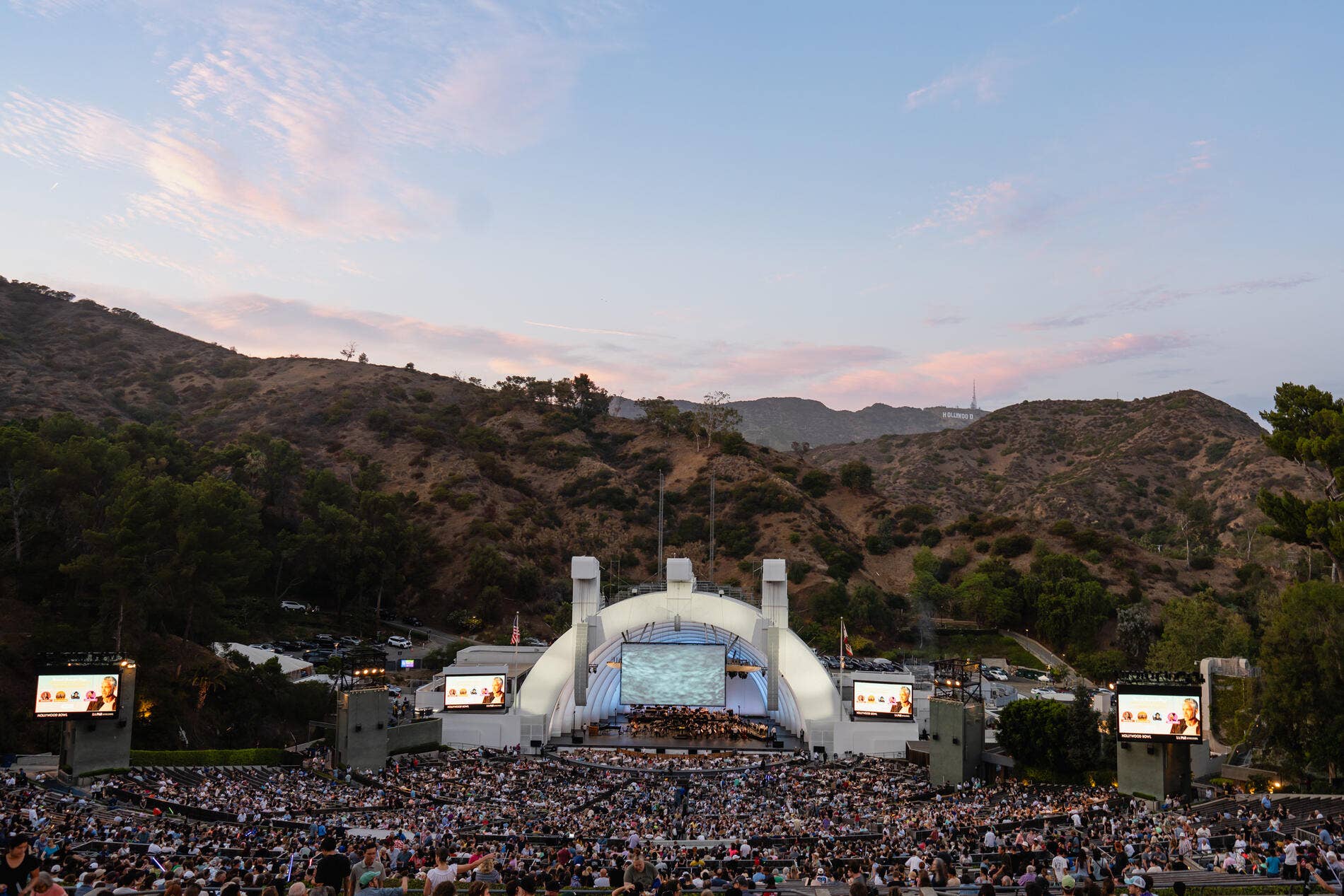 Escenario de un teatro al aire libre con público en el Hollywood Bowl al atardecer, rodeado de colinas y árboles