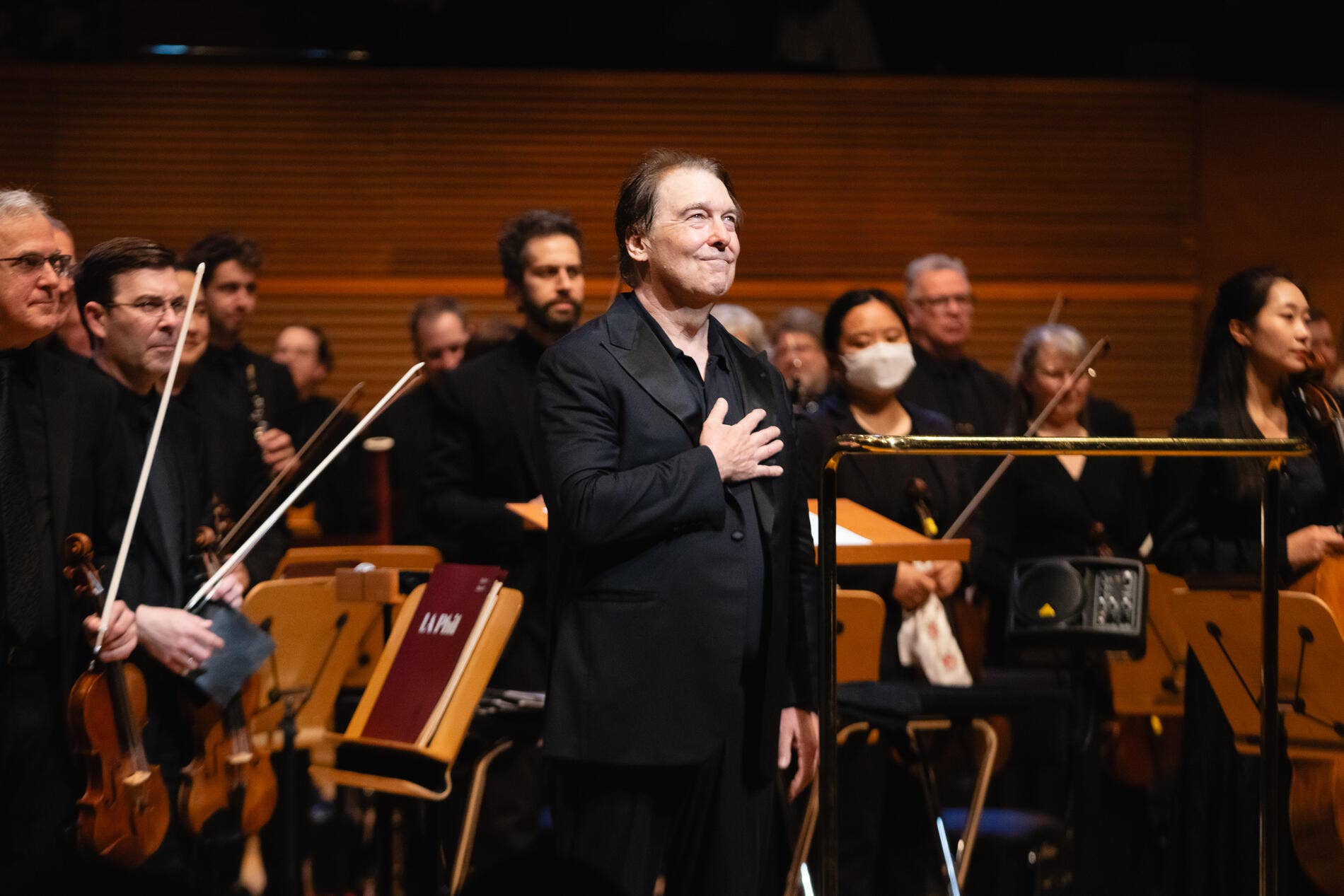 director de orquesta chaqueta negra dirige a la orquesta, con la mano en el pecho, con los músicos y los instrumentos a sus espaldas.