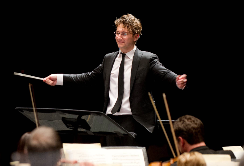 Curly-haired conductor in a suit gestures with both arms while leading an orchestra from a music stand onstage