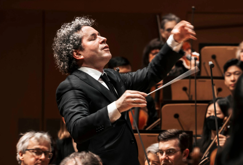 Conductor Gustavo Dudamel in black suit leads an orchestra, arms raised, baton in hand during a live performance.