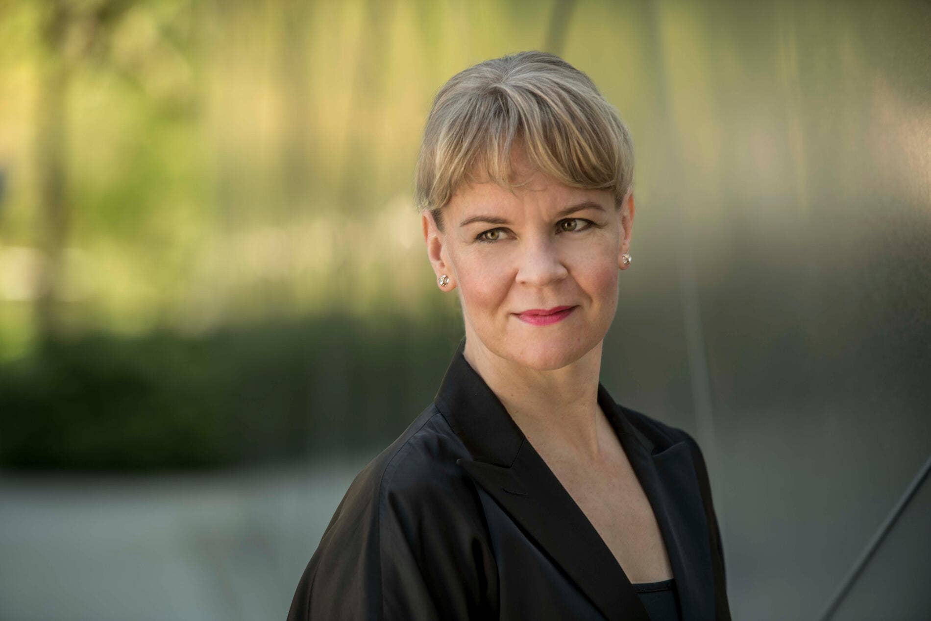 Susanna Mälkki with short gray hair and earrings, wearing a black blazer, against a blurred outdoor background.