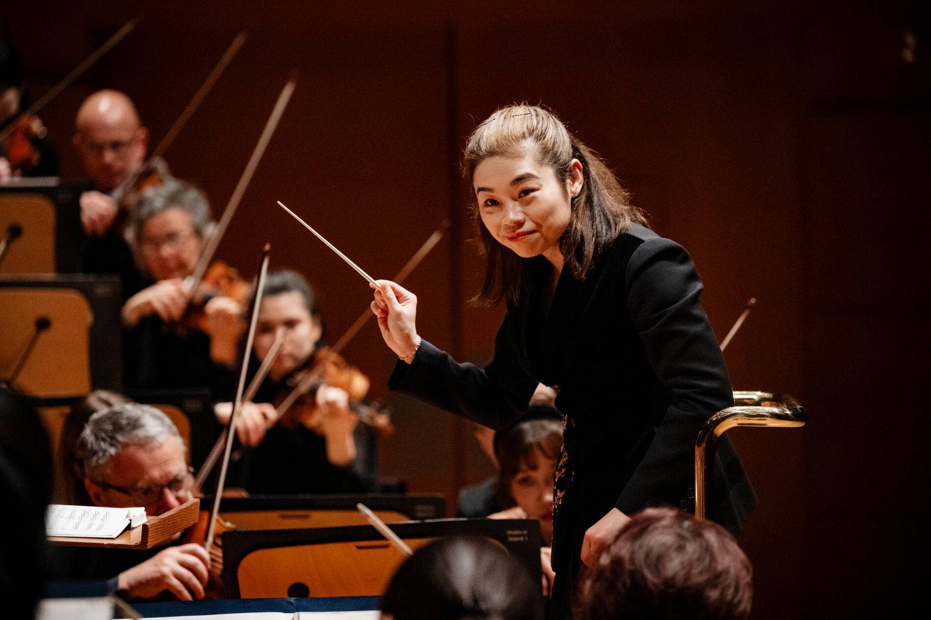 Female conductor, Elim Chan, leads an orchestra, holding a baton as musicians play in a concert hall.