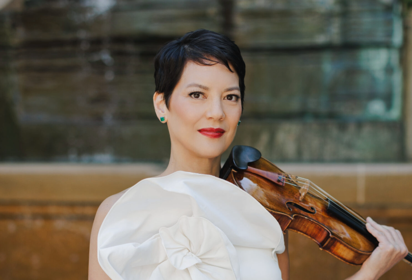 A smiling Anne Akiko Meyers in white holding a violin outdoors with soft-focus background and city wall behind her