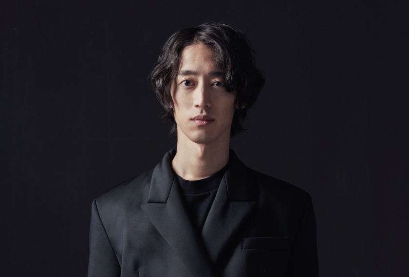Retrato de Hayato Sumino, con el pelo ondulado y vestido con una chaqueta oscura, sobre un fondo oscuro, mirando a la cámara