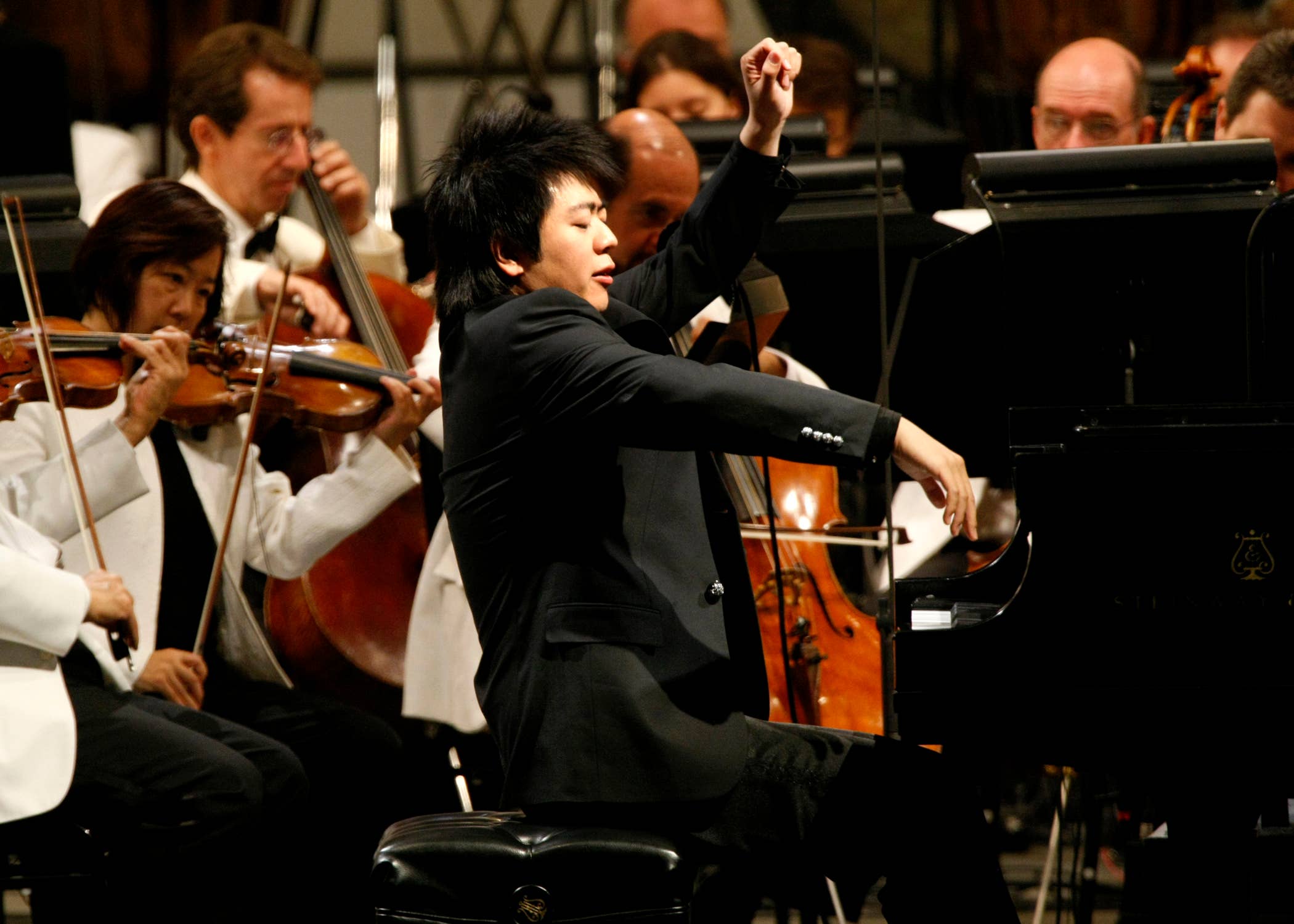 Un pianista en concierto levanta la mano mientras los miembros de la orquesta tocan los instrumentos de cuerda a sus espaldas.