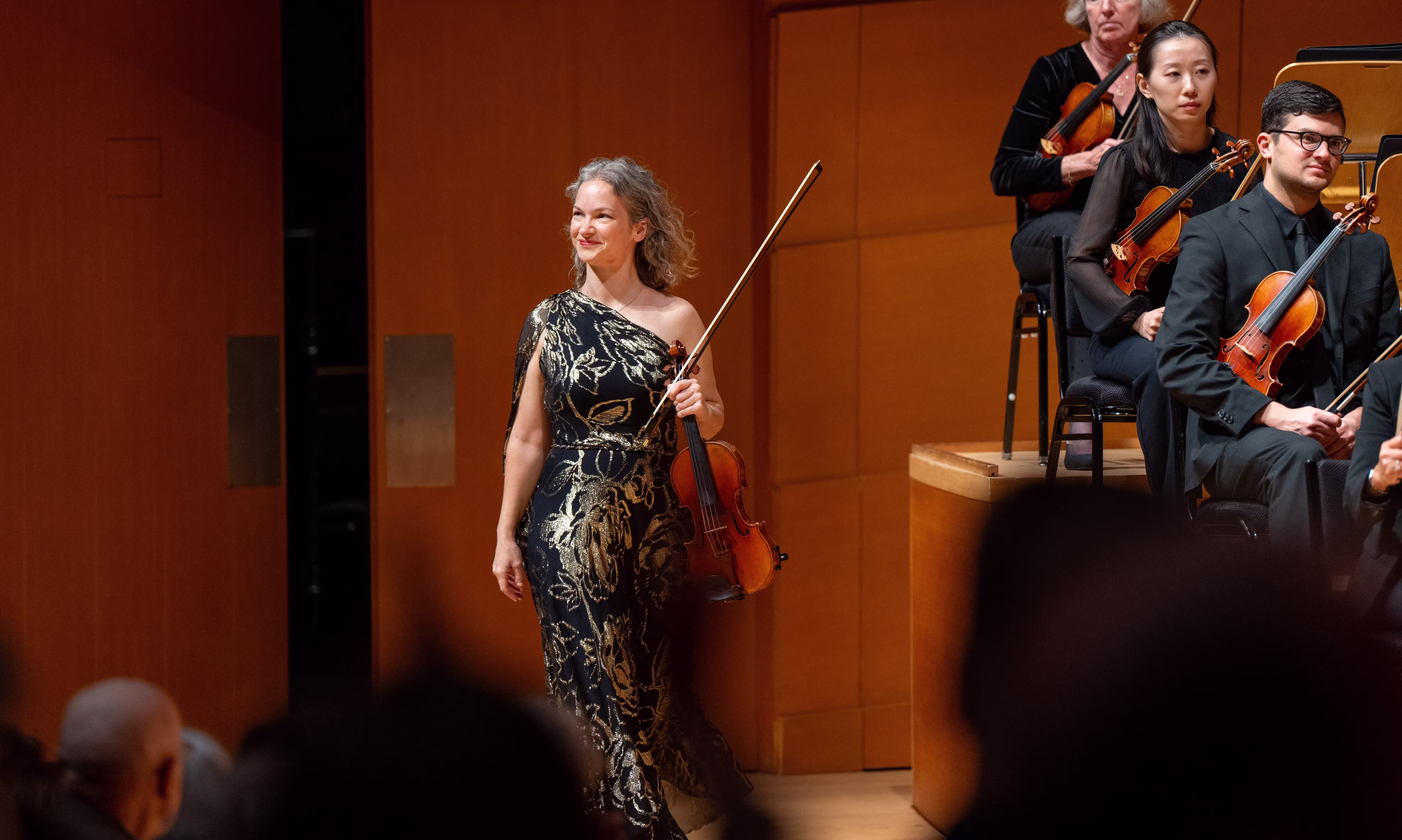 Woman in black and gold gown walks onto stage holding a violin.