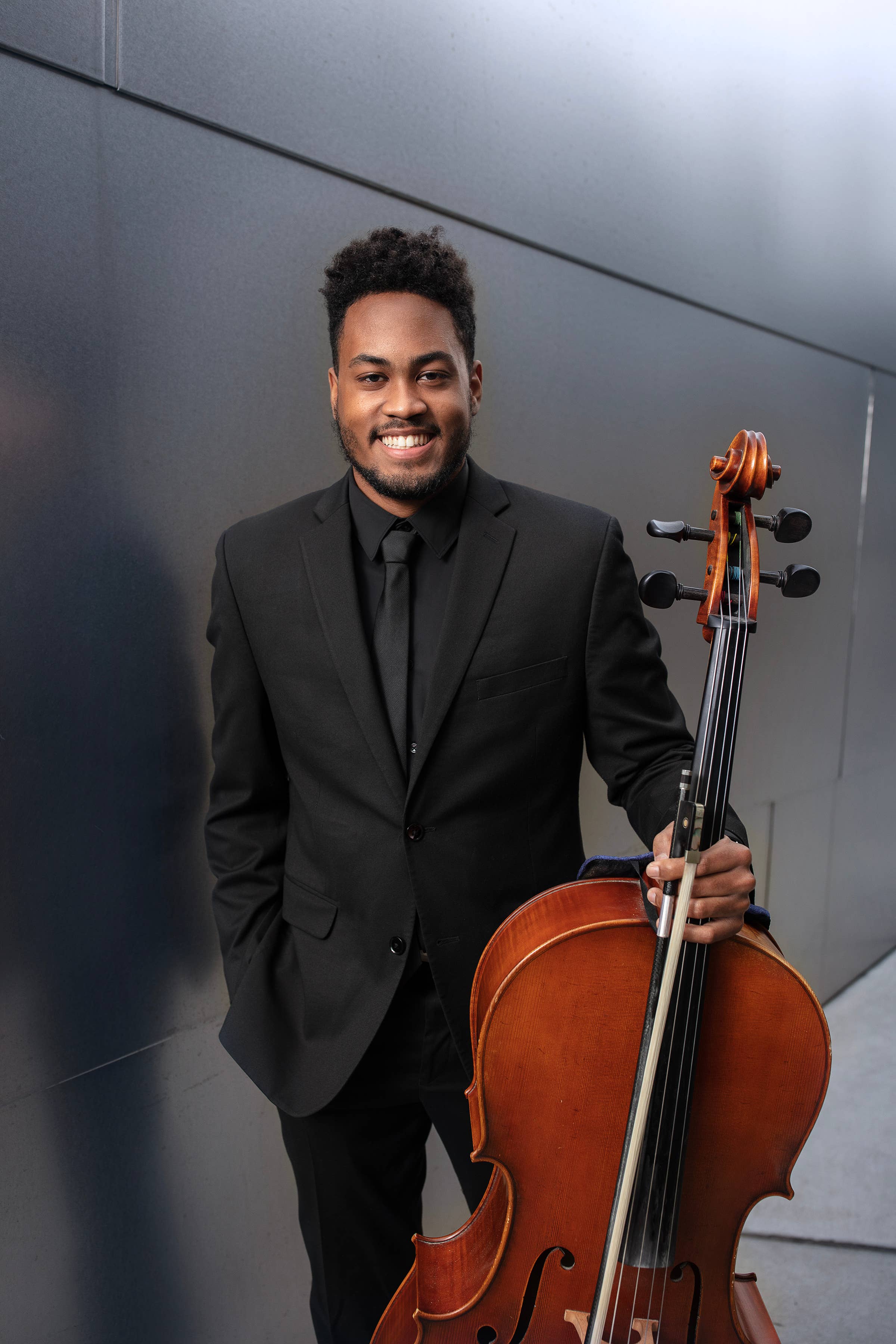 Hombre sonriente con traje negro que sostiene un violonchelo marrón, con una pared gris de fondo.