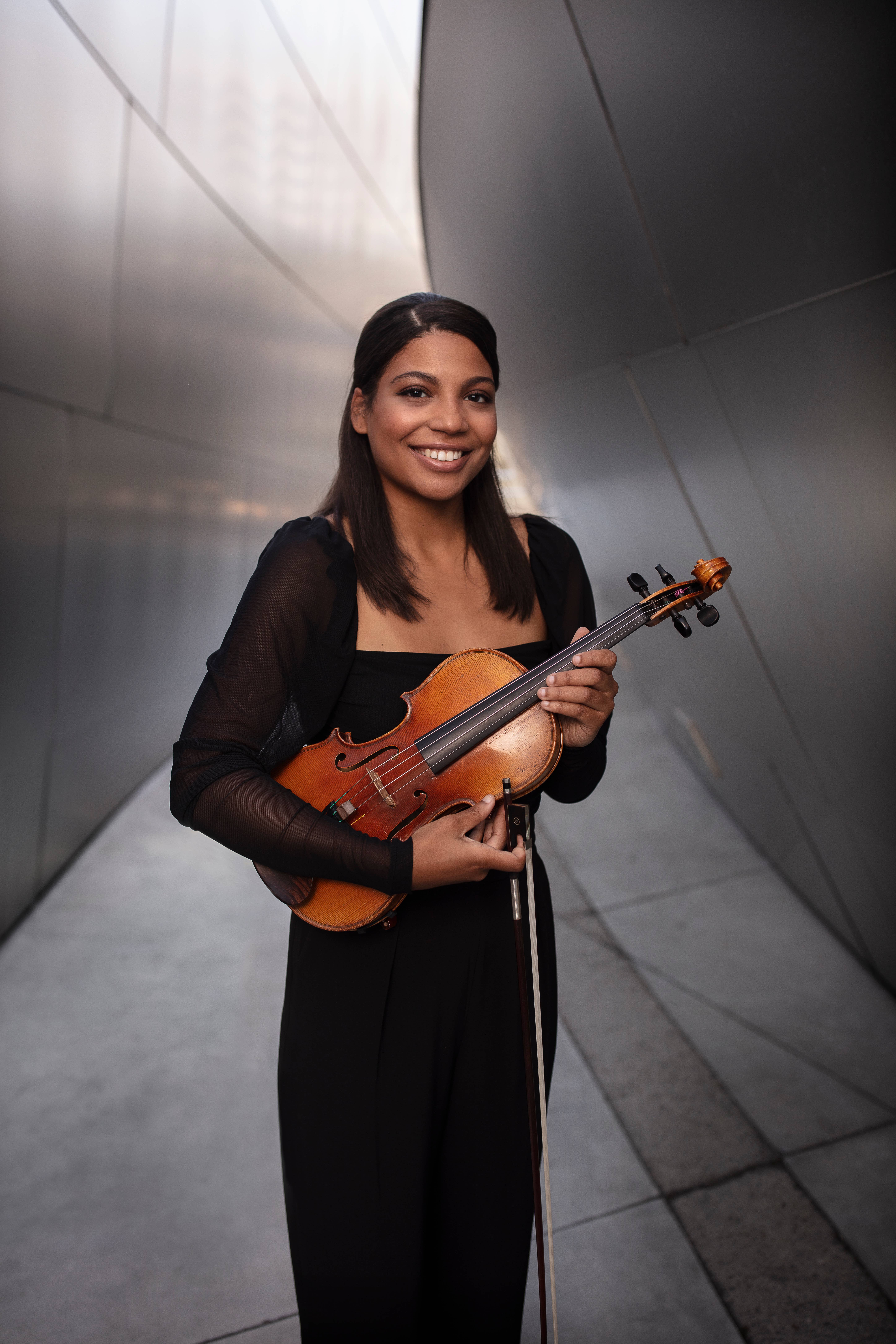 Smiling woman in black holding a violin on a bow stand in a modern tiled hallway.
