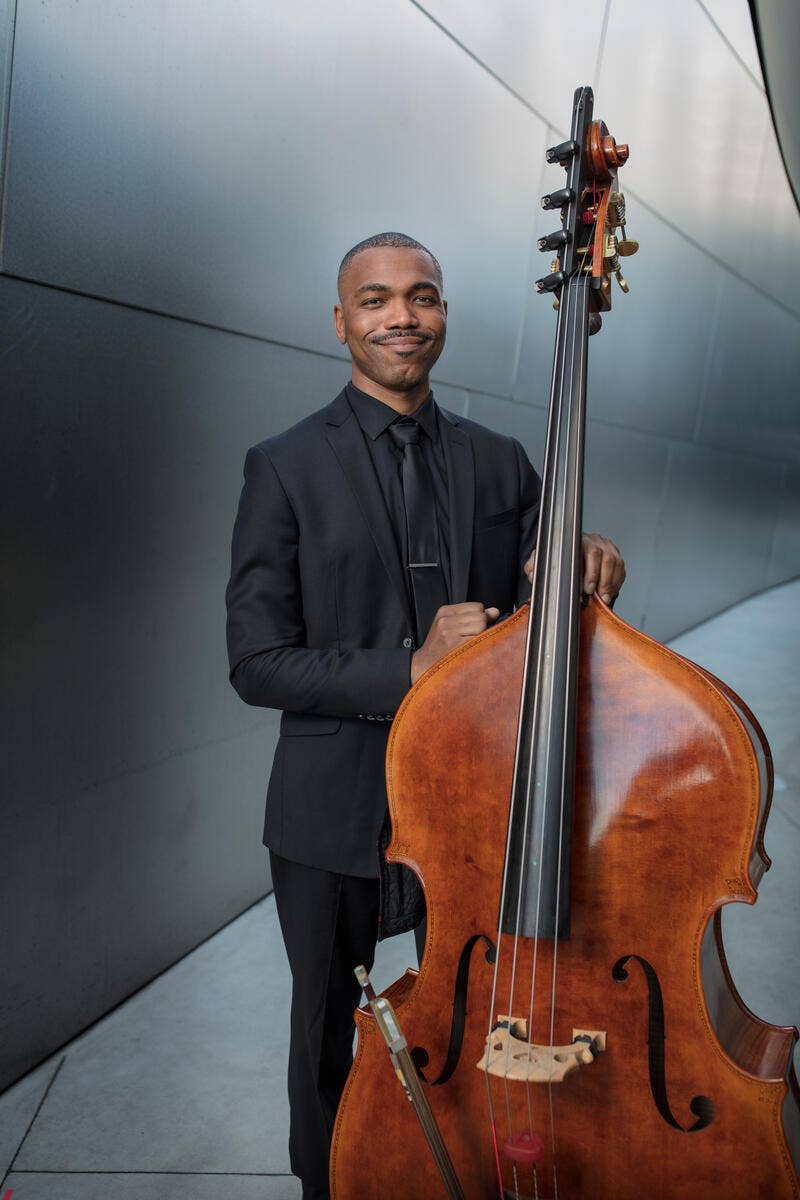 Smiling man in a black suit holding a brown bass, standing against a modern blue wall with diagonal lines