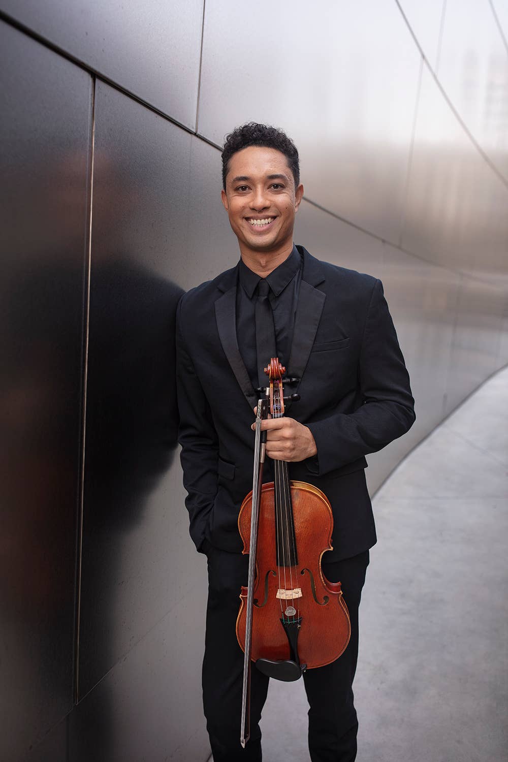 Smiling musician in a black suit holding a wooden violin indoors against a modern curved wall.
