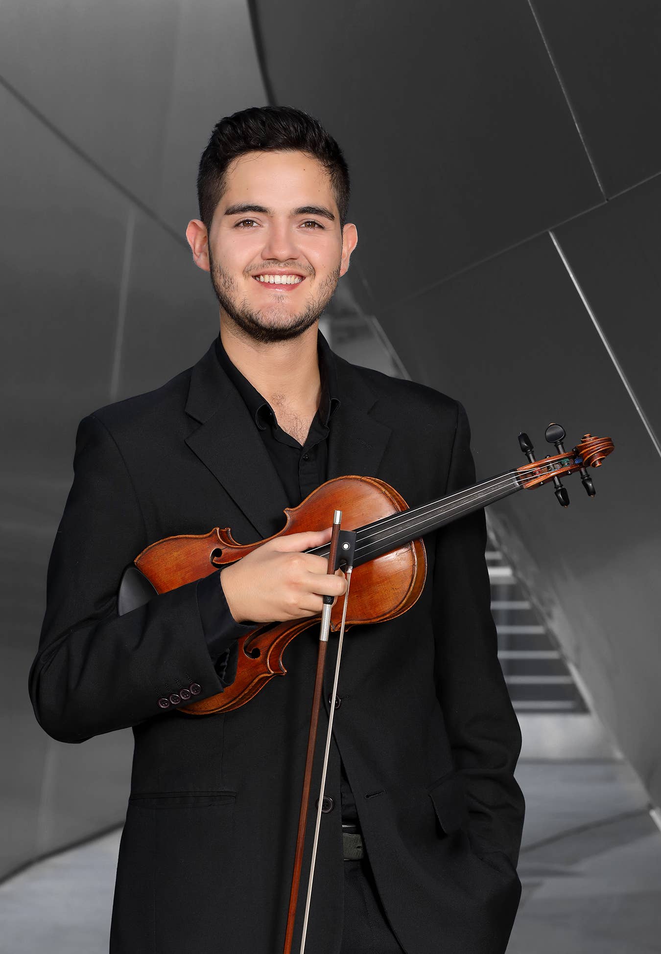 Hombre sonriente con traje oscuro que sostiene un violín, posando con una moderna escalera gris como fondo.