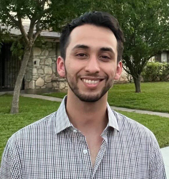Smiling young man with short dark hair and trimmed beard outdoors in a green garden.