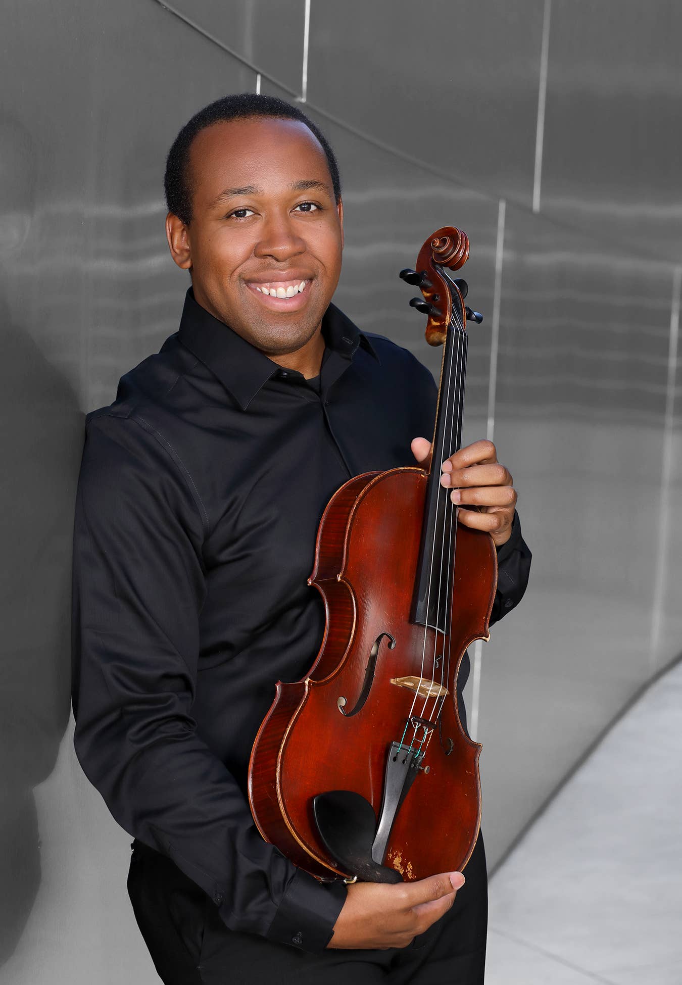 Un hombre sonriente con una camiseta negra que sostiene un violín marrón delante de un fondo gris con paneles de luz verticales