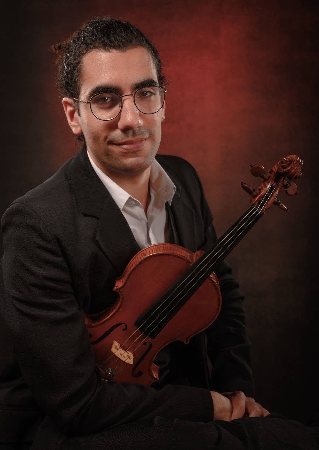 Smiling man in suit with glasses holding a brown violin against a dark red studio backdrop.