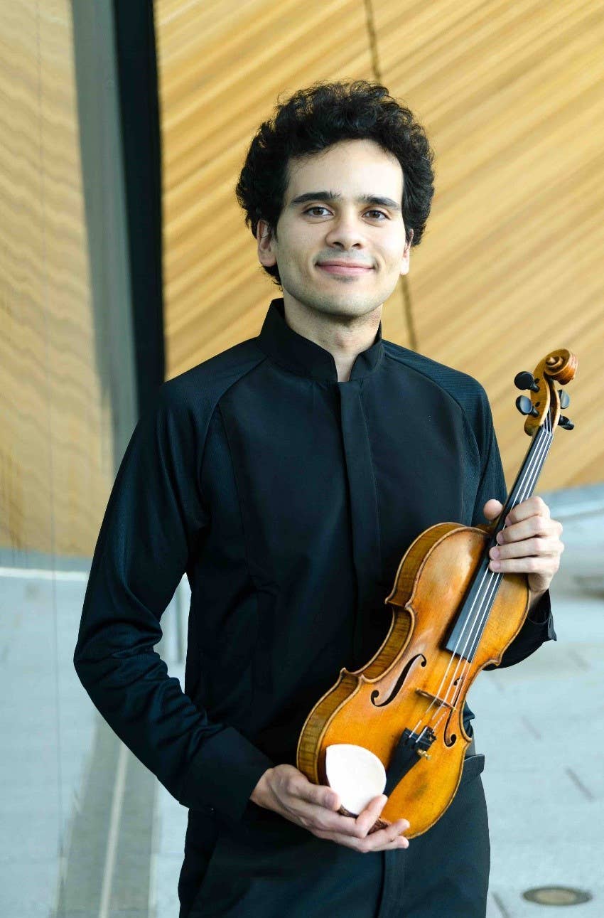Young man in black holding a brown violin, smiling in front of a light beige wall.