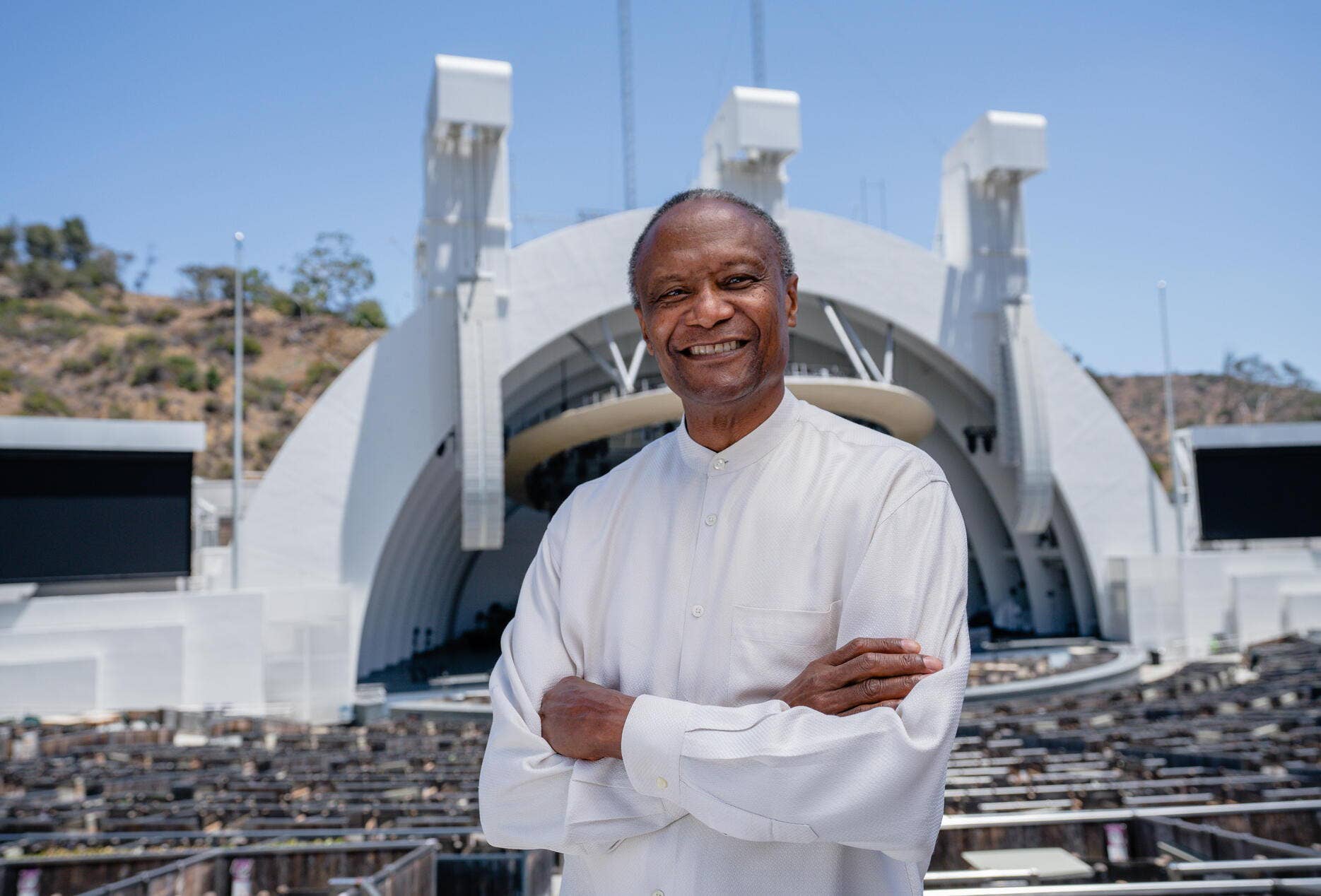 Un hombre sonriente con camisa blanca, de pie frente a un estadio moderno con altos arcos blancos bajo un cielo azul