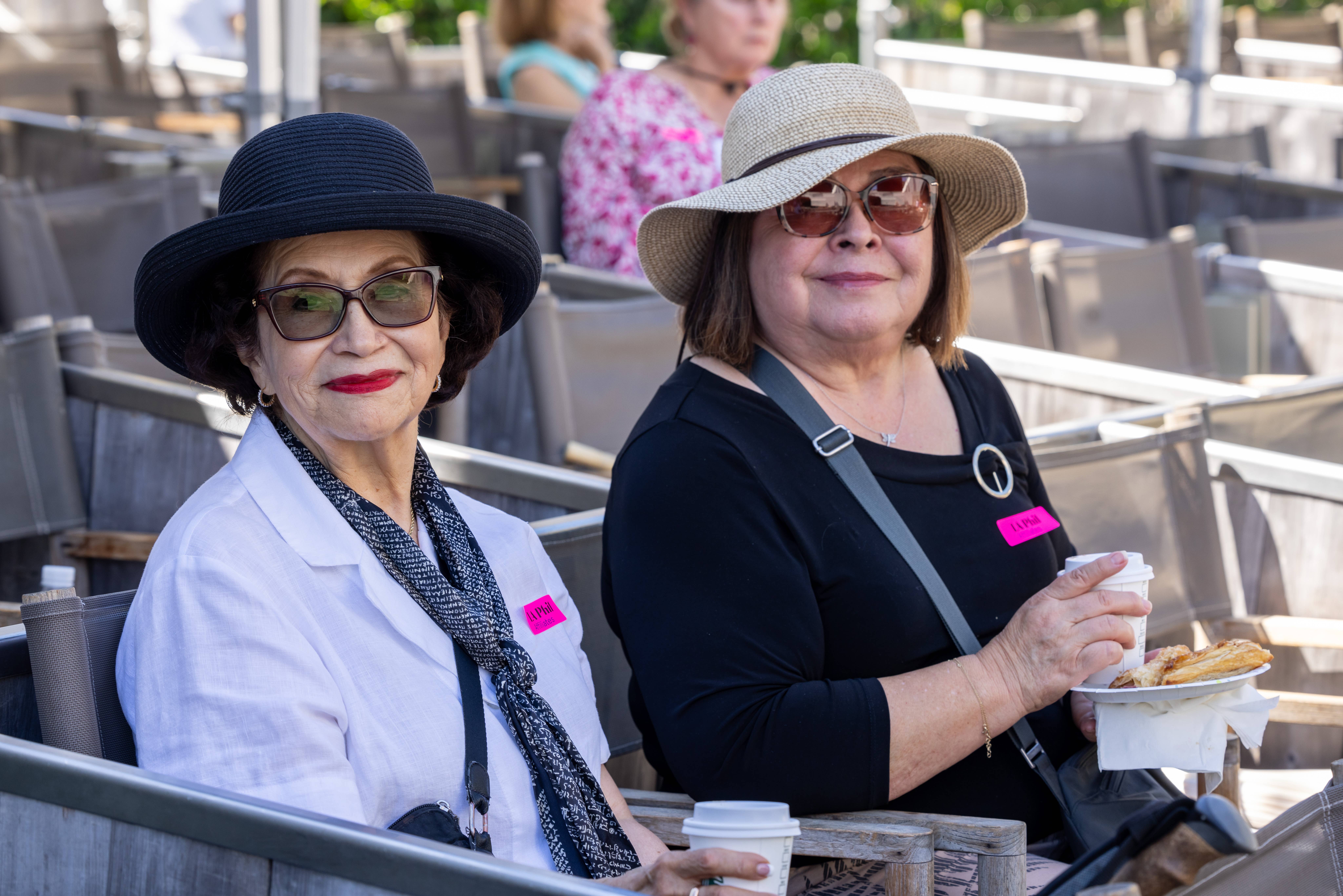 Dos mujeres sentadas en la terraza de una cafetería, con sombreros de ala ancha, gafas de sol y etiquetas con sus nombres, sosteniendo café y comida.