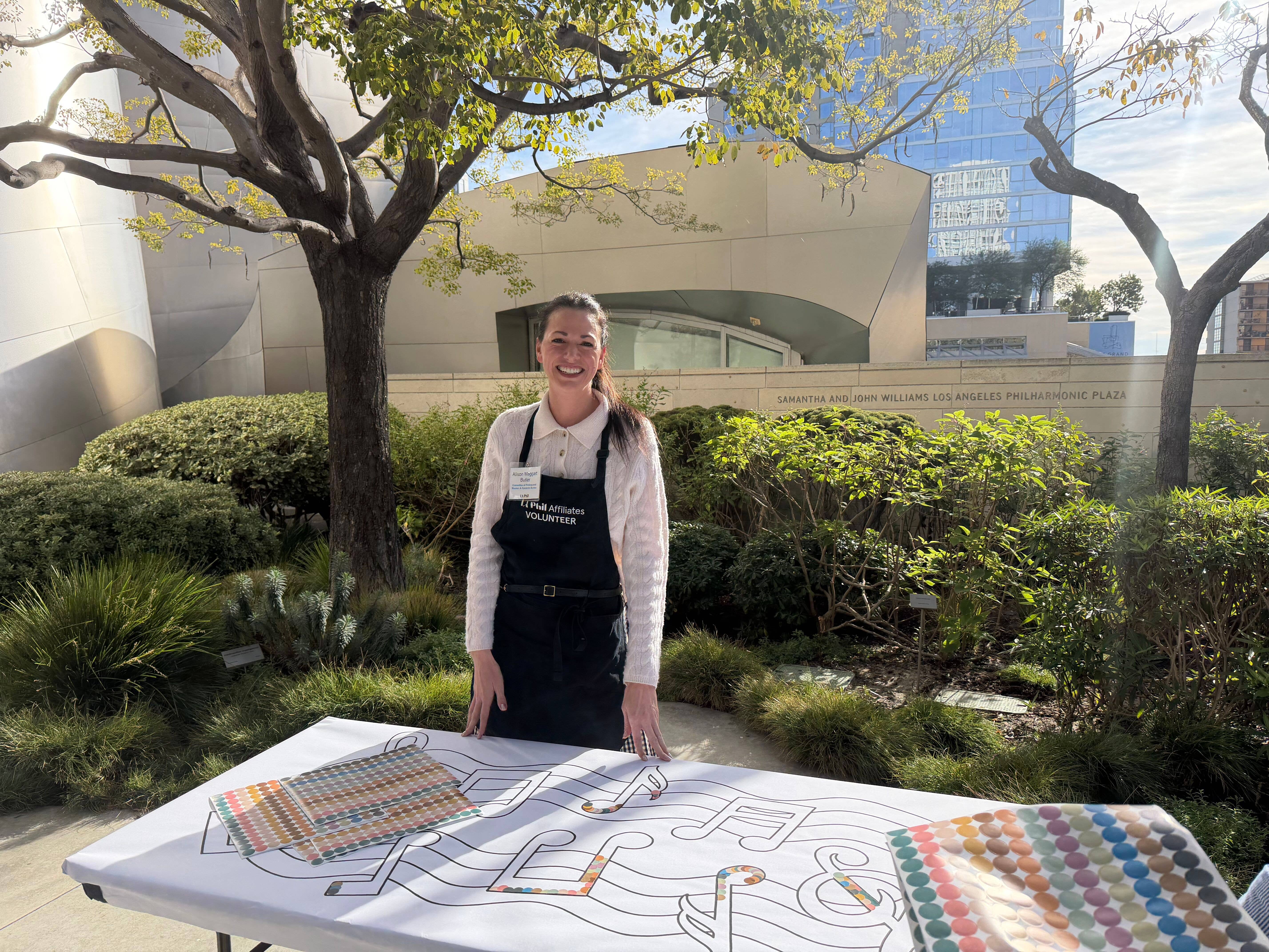 Un voluntario sonriente, con delantal, está junto a una mesa con material del evento, en una plaza ajardinada con grandes edificios al fondo.