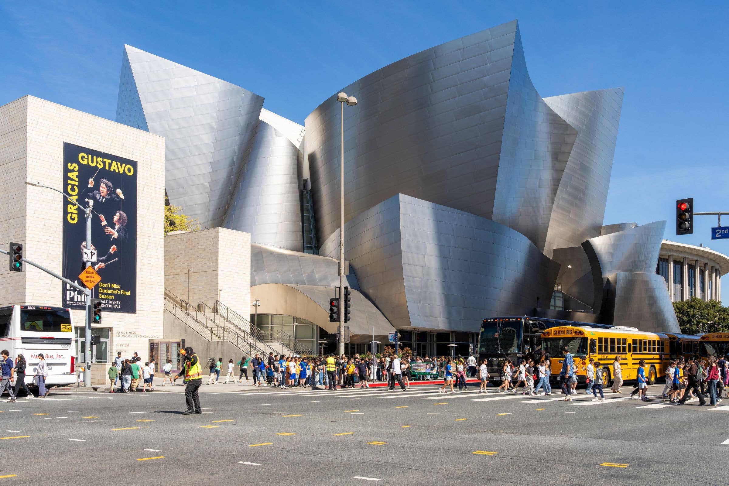 Crowd gathers by a modern concert hall with silver curved facade, traffic lights, and school buses on a sunny day