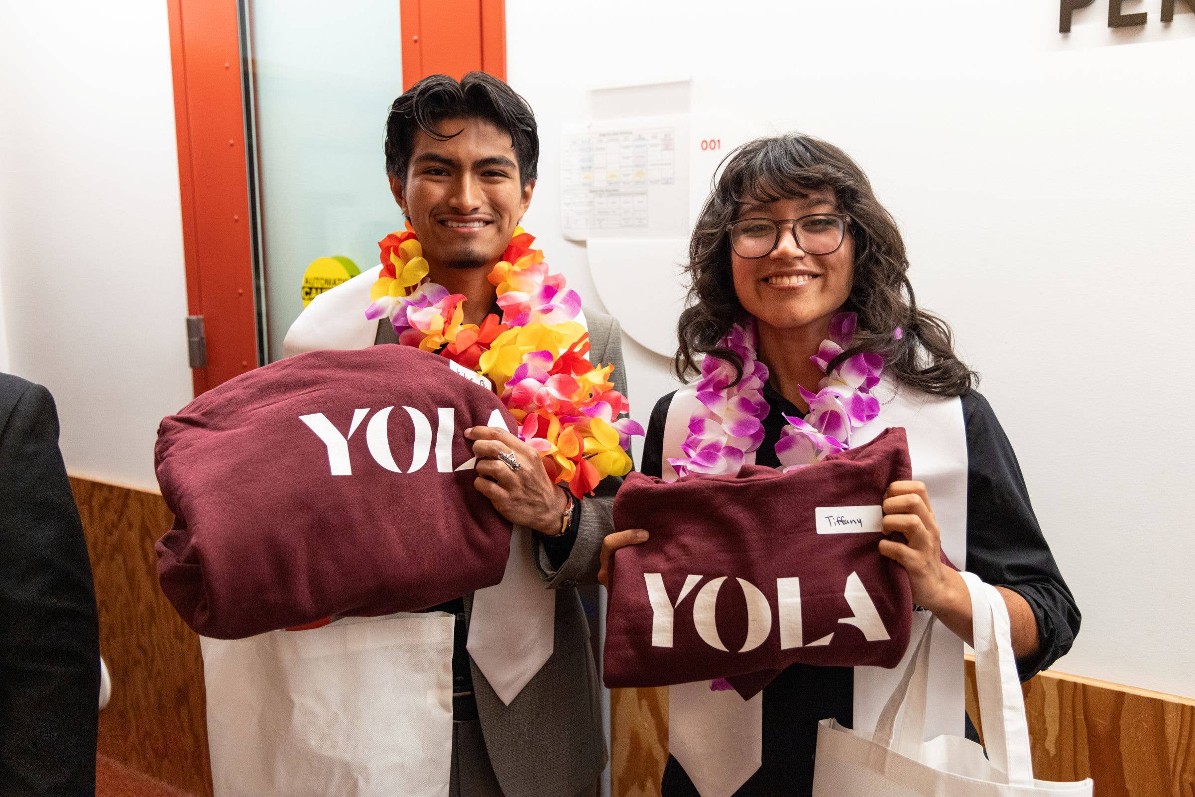 Two students smiling while holding maroon YO!LA and garlands of colorful flowers indoors