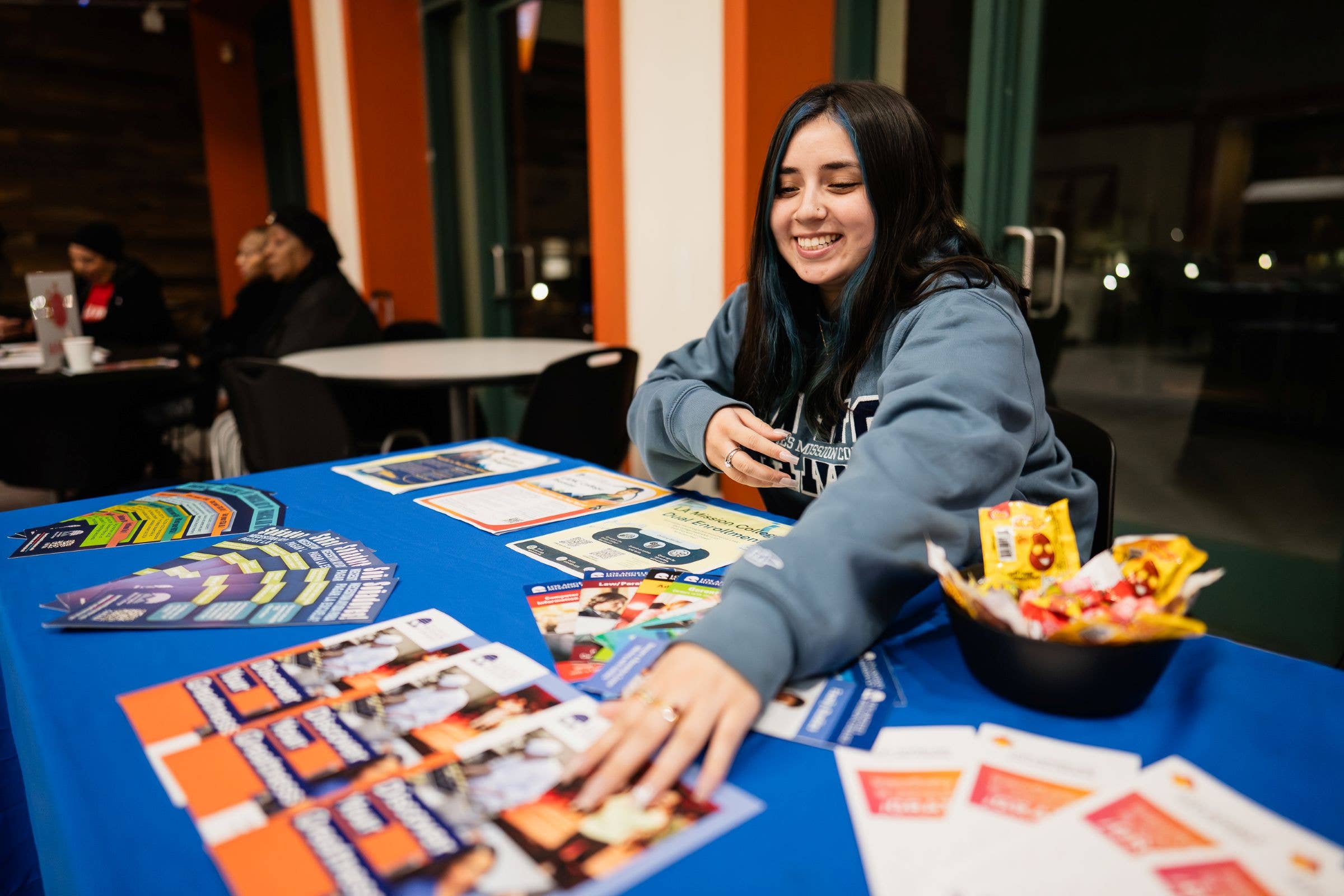 Smiling woman at an event table arranging colorful brochures, with another person seated in the background.