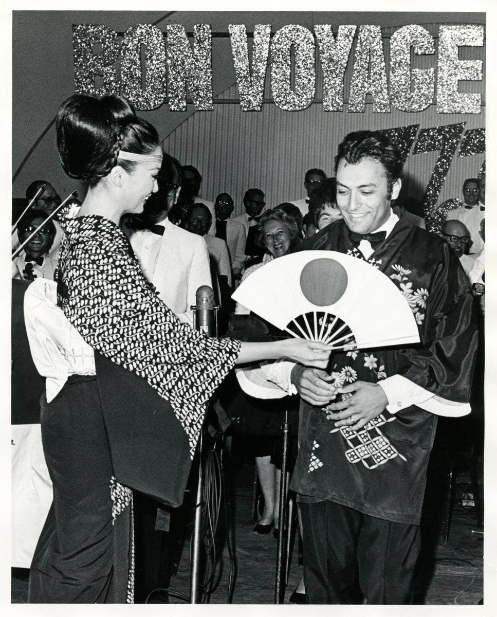 Woman and man onstage exchange a decorative fan and bow as a large “CONGRATULATIONS” banner hangs behind them