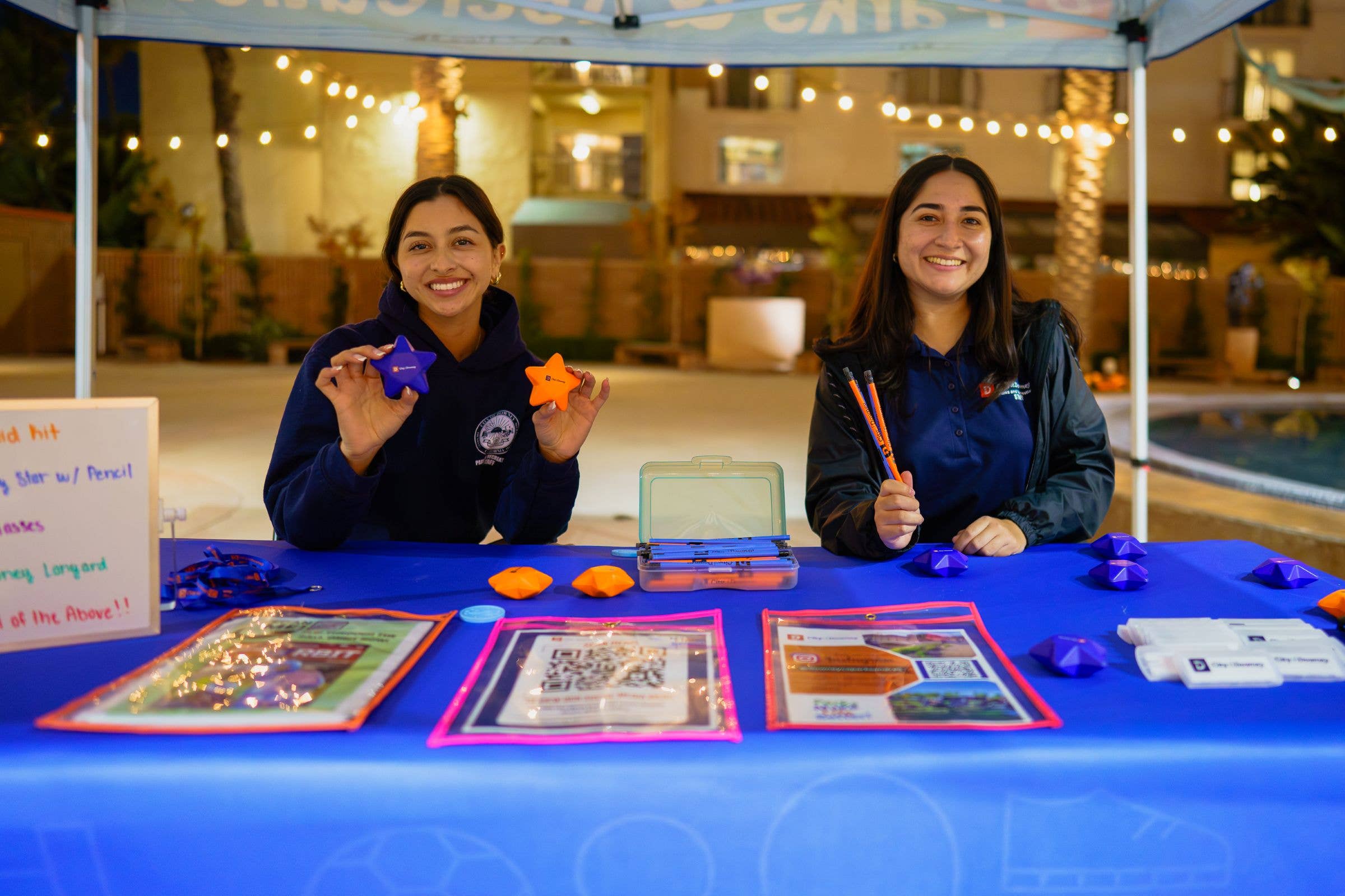 Two women at an outdoor booth holding colorful star-shaped items under a canopy
