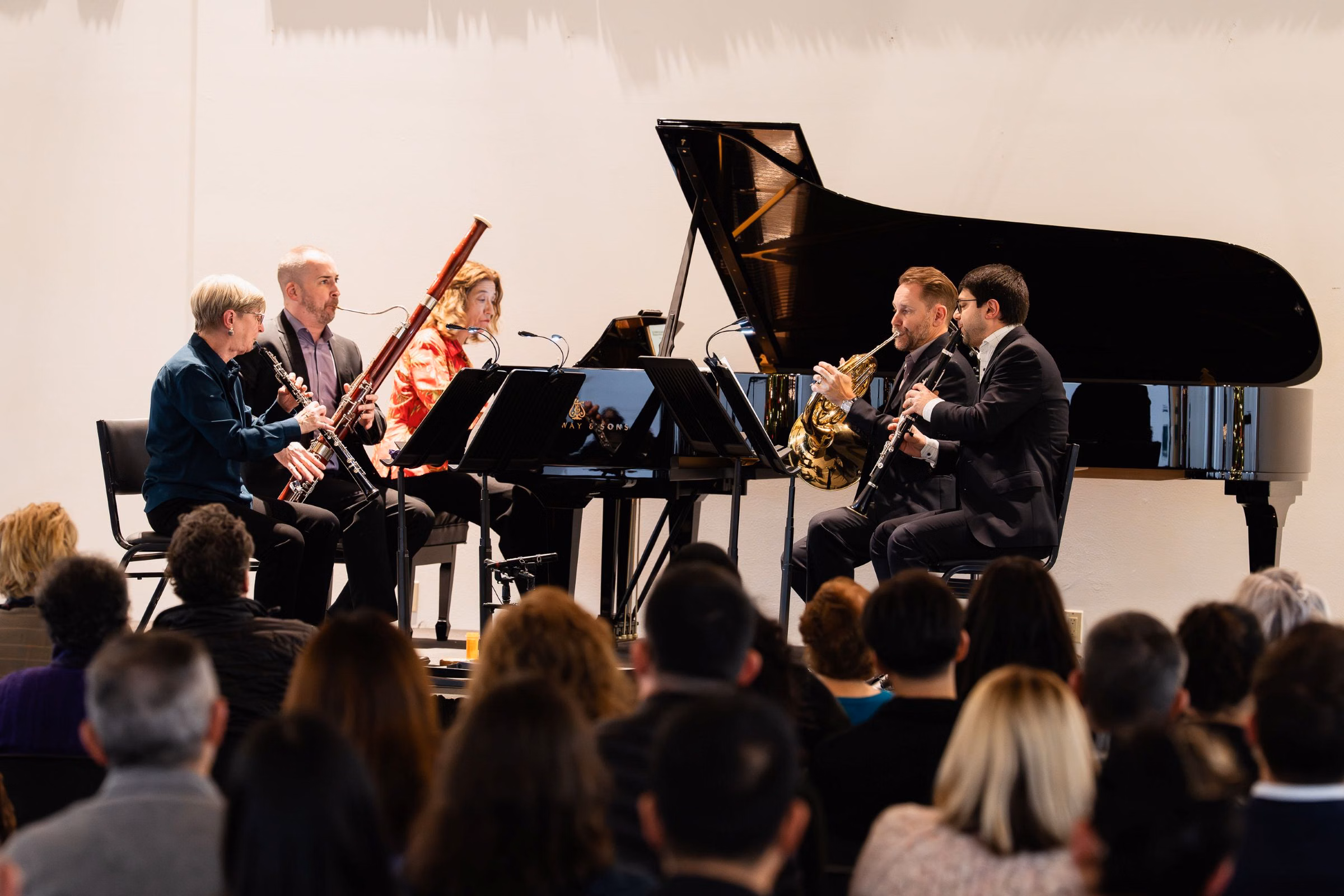 Orchestra musicians play clarinet, flute, and trumpet beside a grand piano at a concert, with audience in foreground