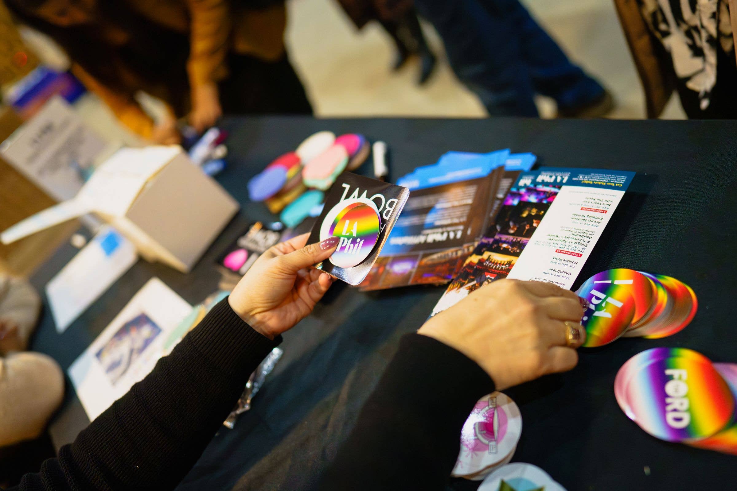 Hands holding circular festival badges over a table with printed flyers and colorful event cards.