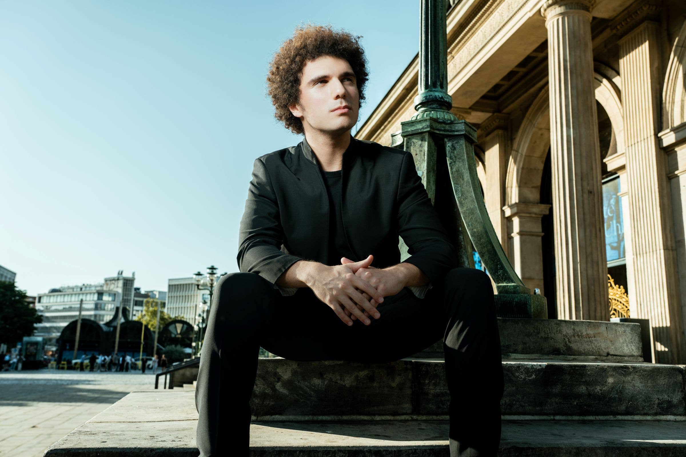 Curly-haired young man in a black jacket sitting beside historic columns on a sunny city plaza