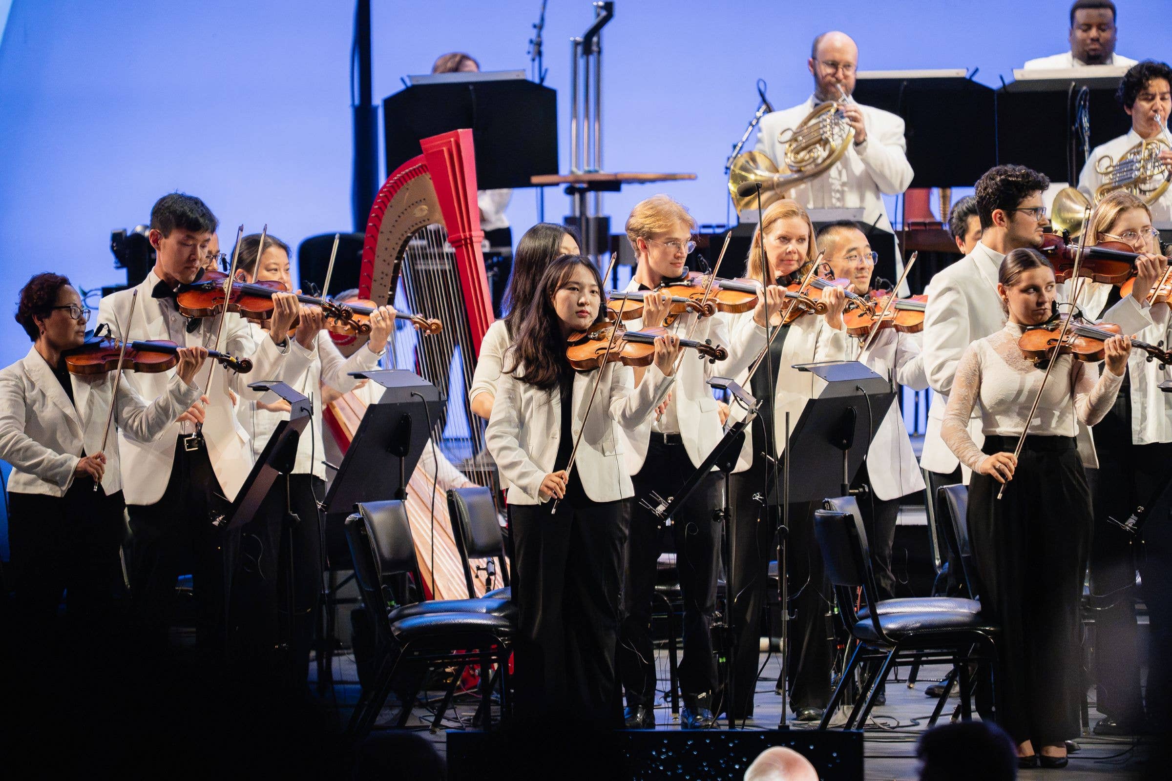 Symphony orchestra in white attire performing with violin and brass on stage under blue lighting