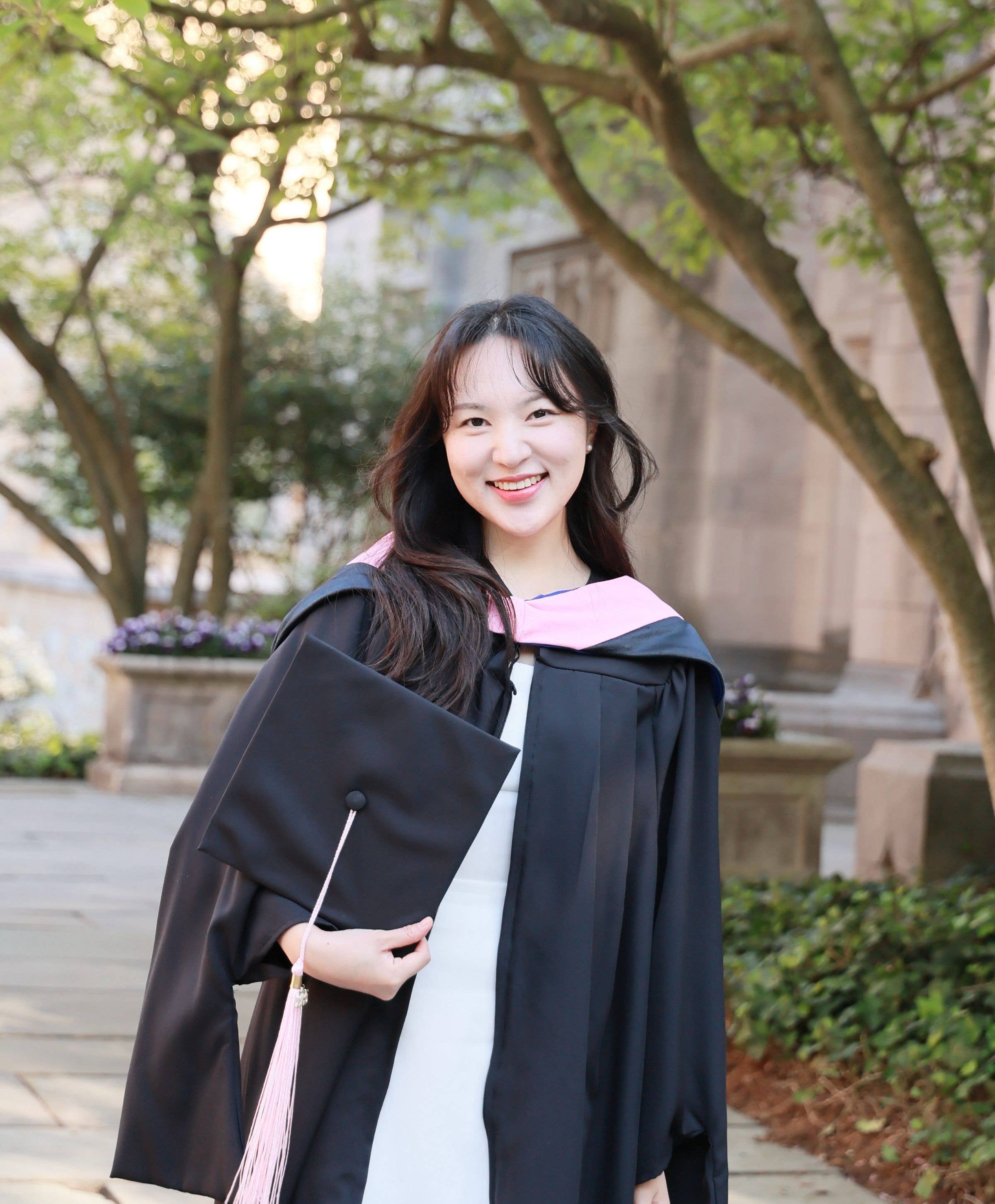 Smiling graduate in black cap and gown outdoors, holding a pink tassel; trees and a stone building in soft focus.