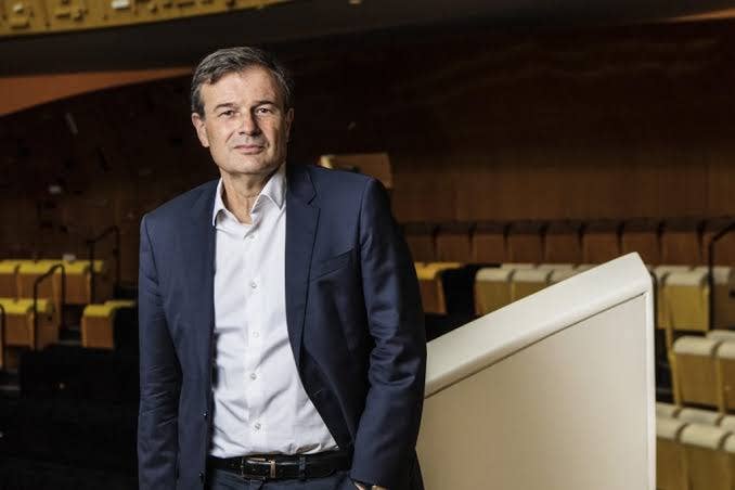 Olivier Mantei dressed in a navy blazer stands beside a white screen in an auditorium with rows of empty seats.