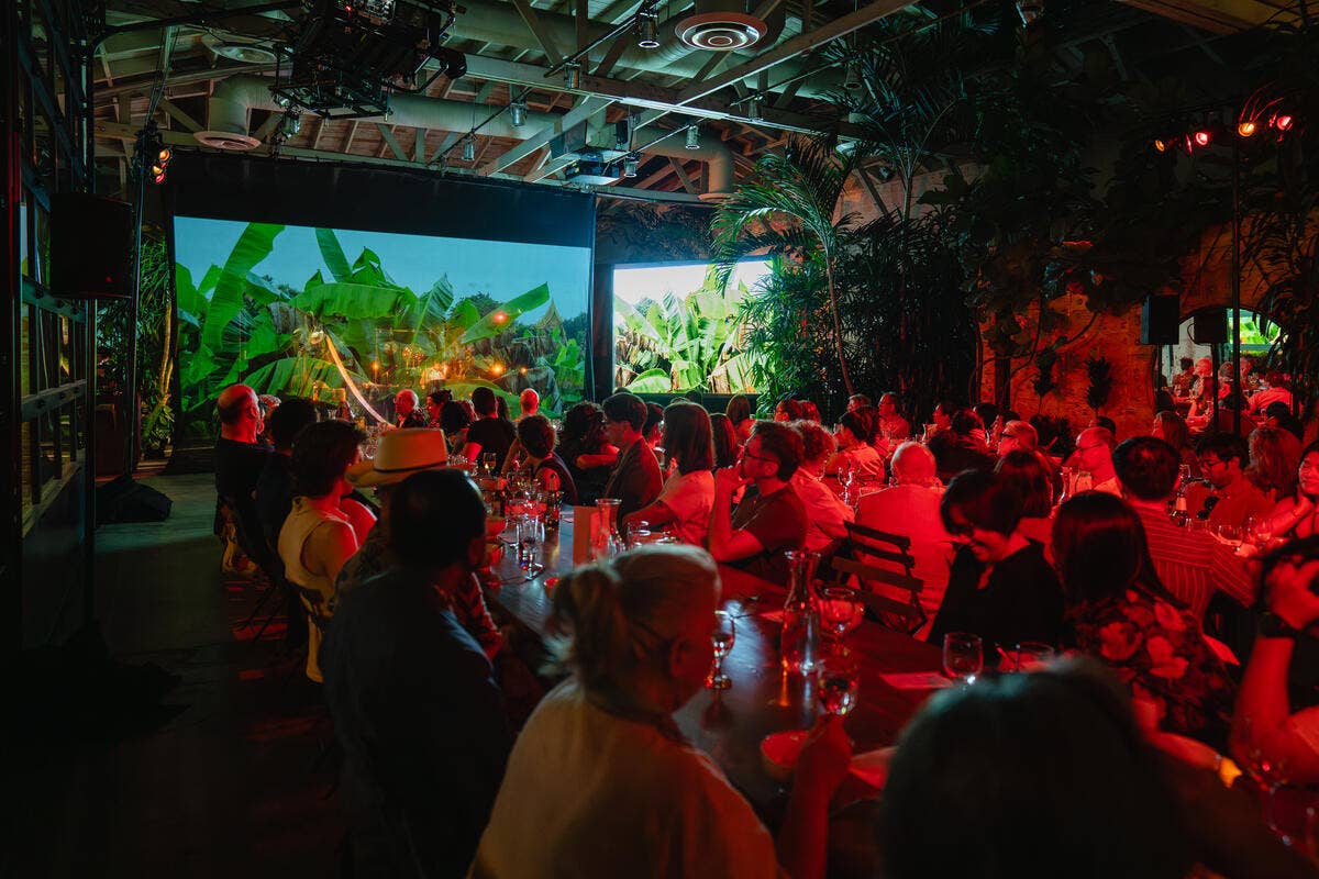 Concert-goers sit in long rows of dining tables with stage lights, watching a projection screen of palm trees.