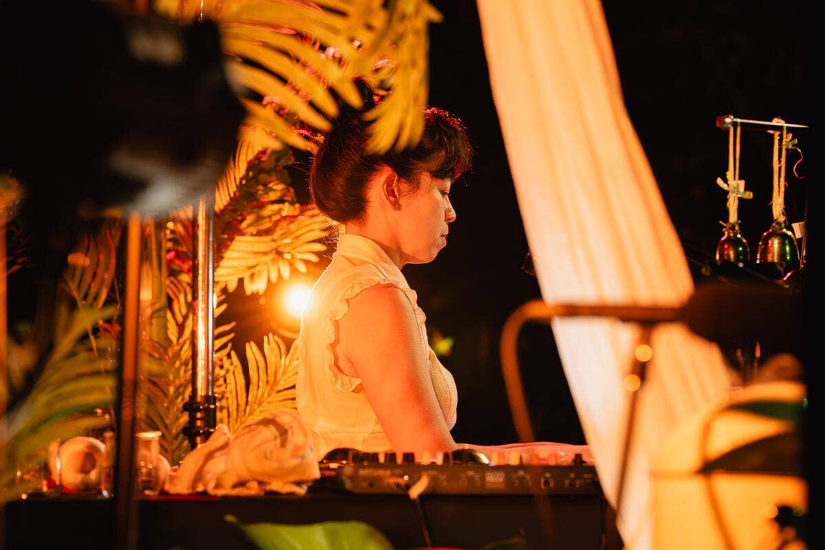 A female musician in white blouse performing with warm lighting in a dark room, surrounded by plants.