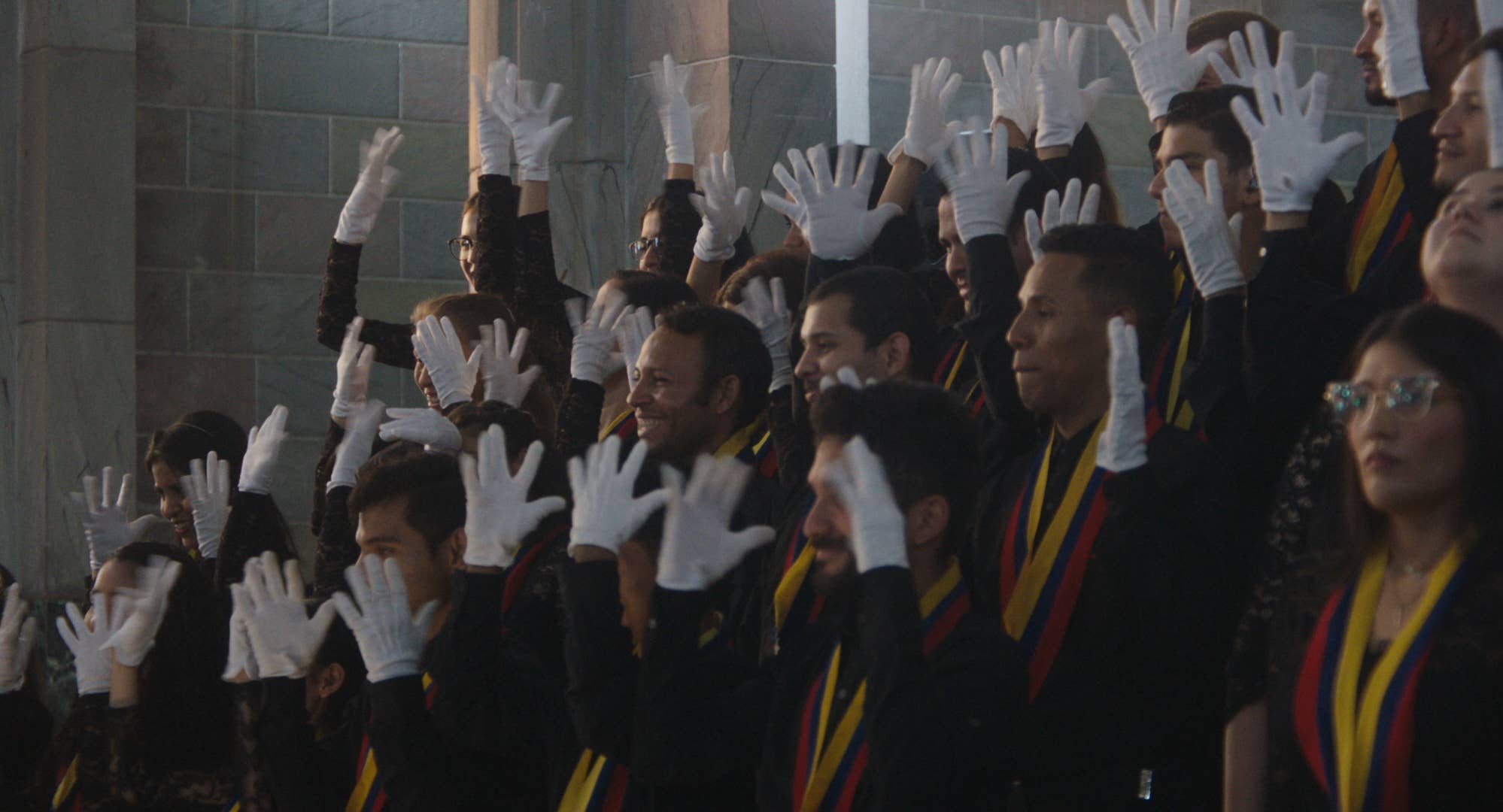 Choir in black-and-yellow uniforms perform with sign language, wearing white gloves, in front of a gray wall.