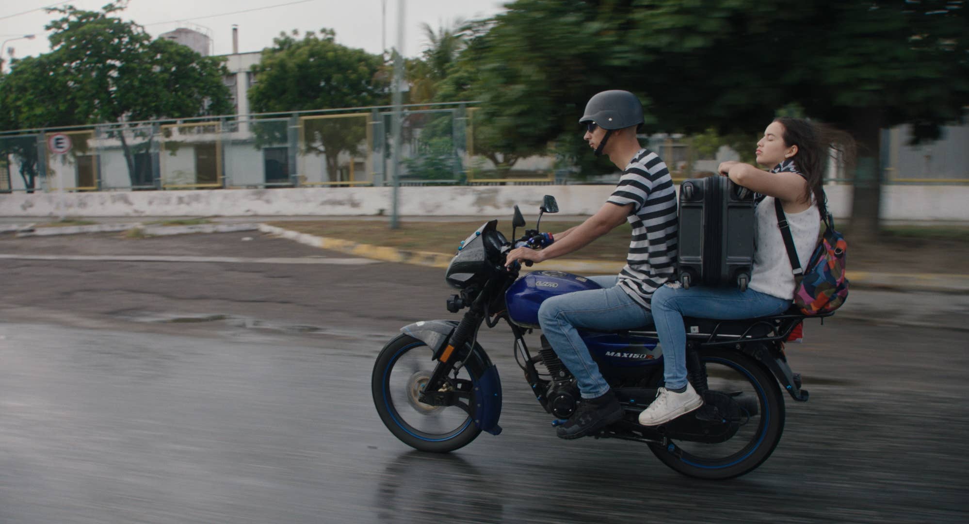 Two riders wearing helmets travel on a blue motorcycle along a road.