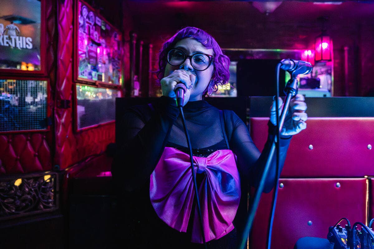 Singer with curly hair and glasses performs at a purple neon-lit bar with a microphone and bright stage lights.