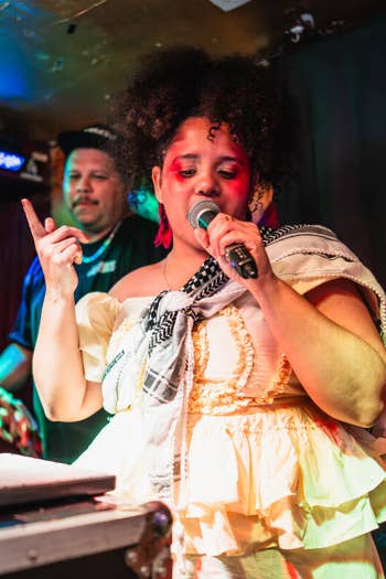Female singer with curly hair in a white dress signs into a microphone under red and green stage lights.