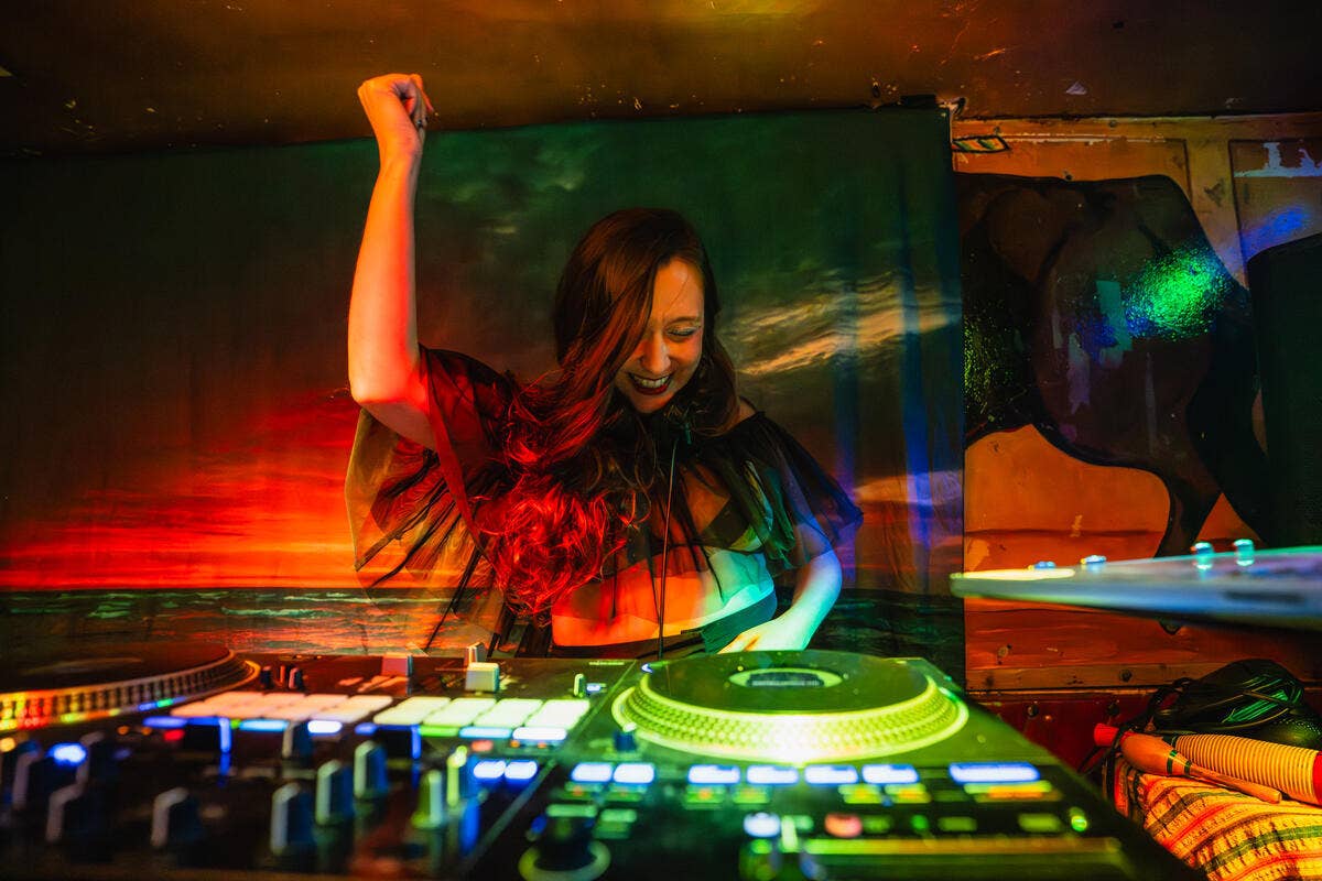 Female DJ raises her left arm in a fist in front of mixing equipment, under red and green stage lights.