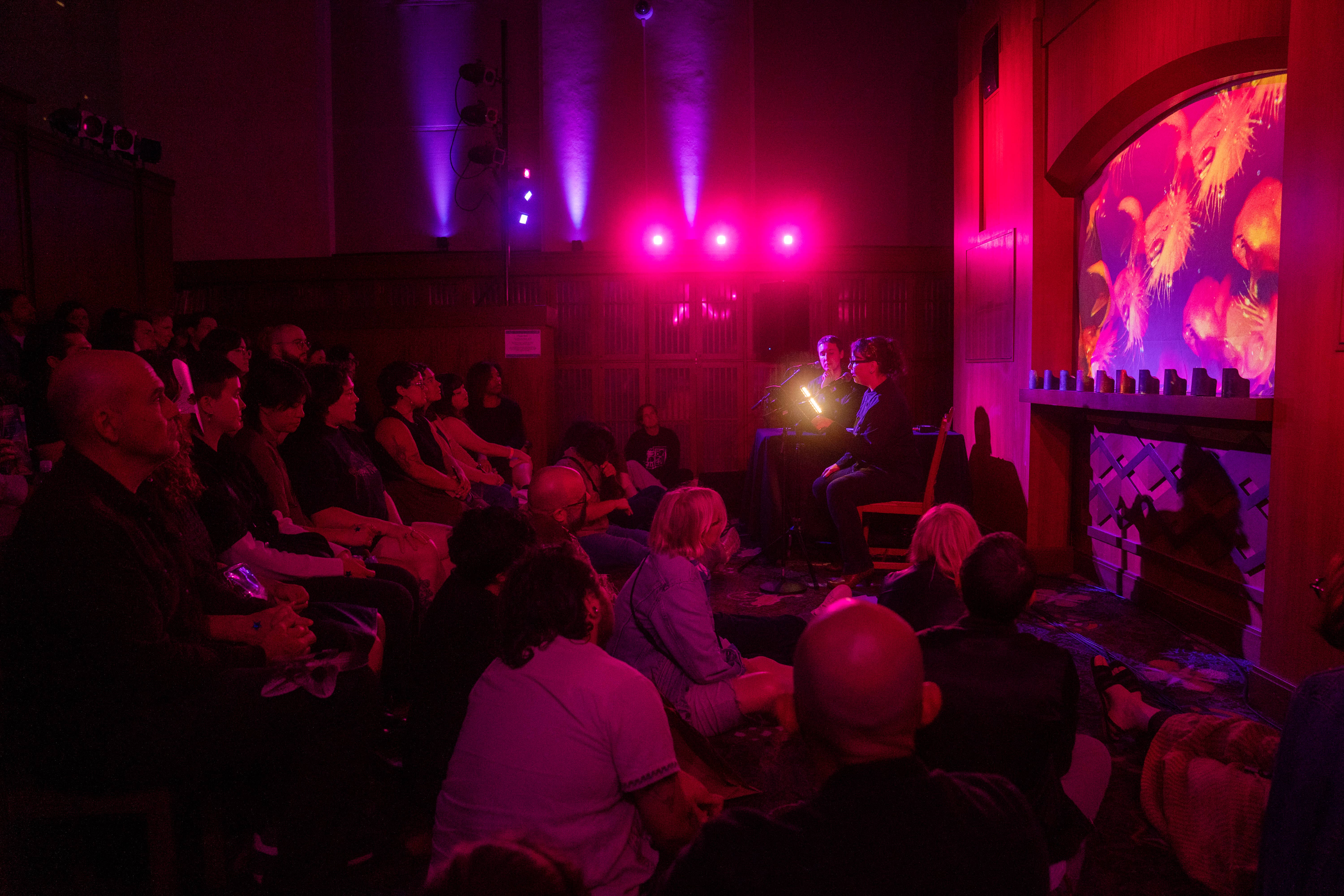 A dark library room crowded with participants lit with pink and purple lights
