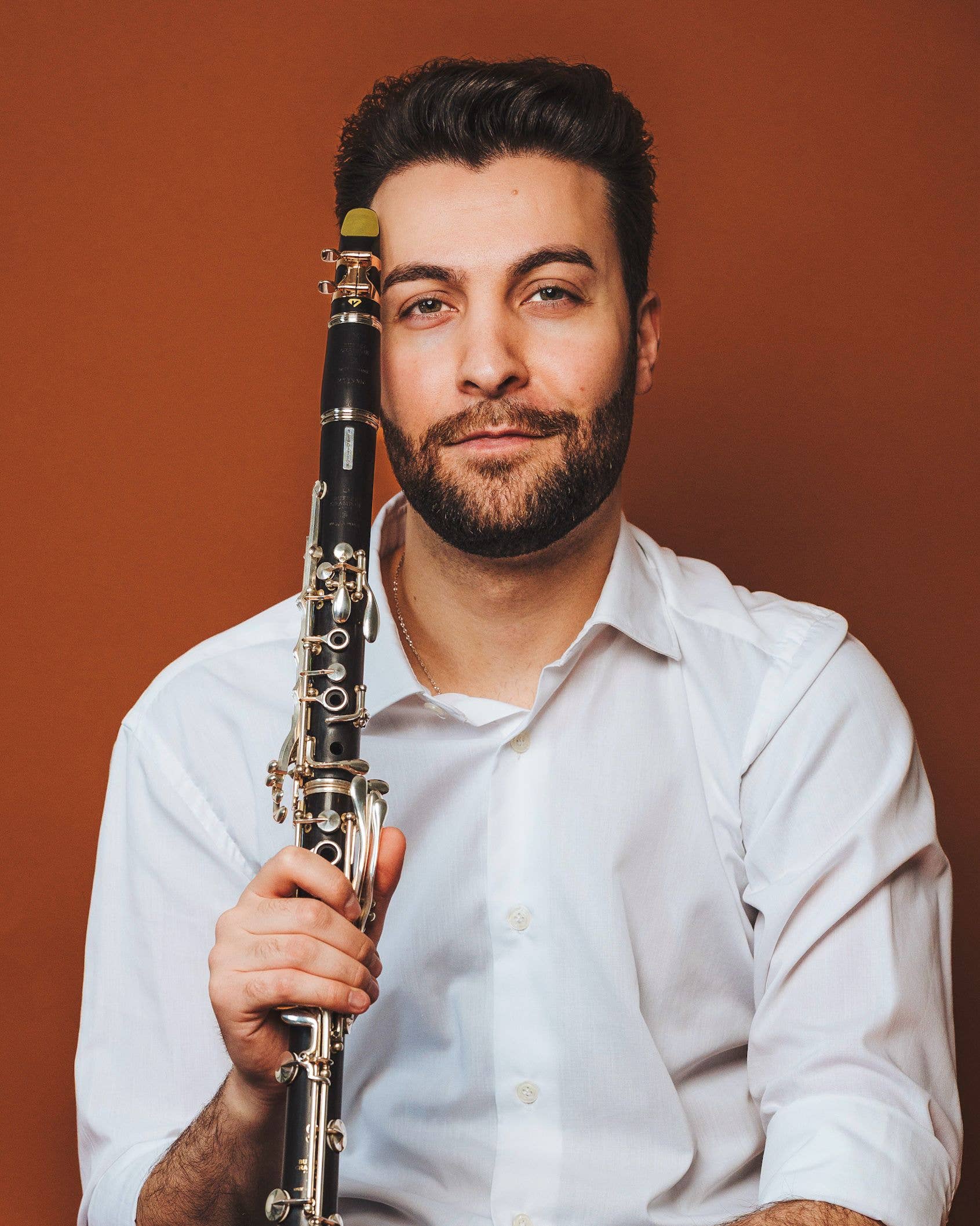Man holding a black clarinet with silver keys, wearing a white shirt against a warm orange background.
