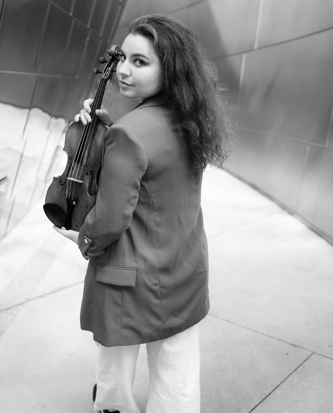 Woman in a blazer holding a violin, standing in a bright tiled hallway.