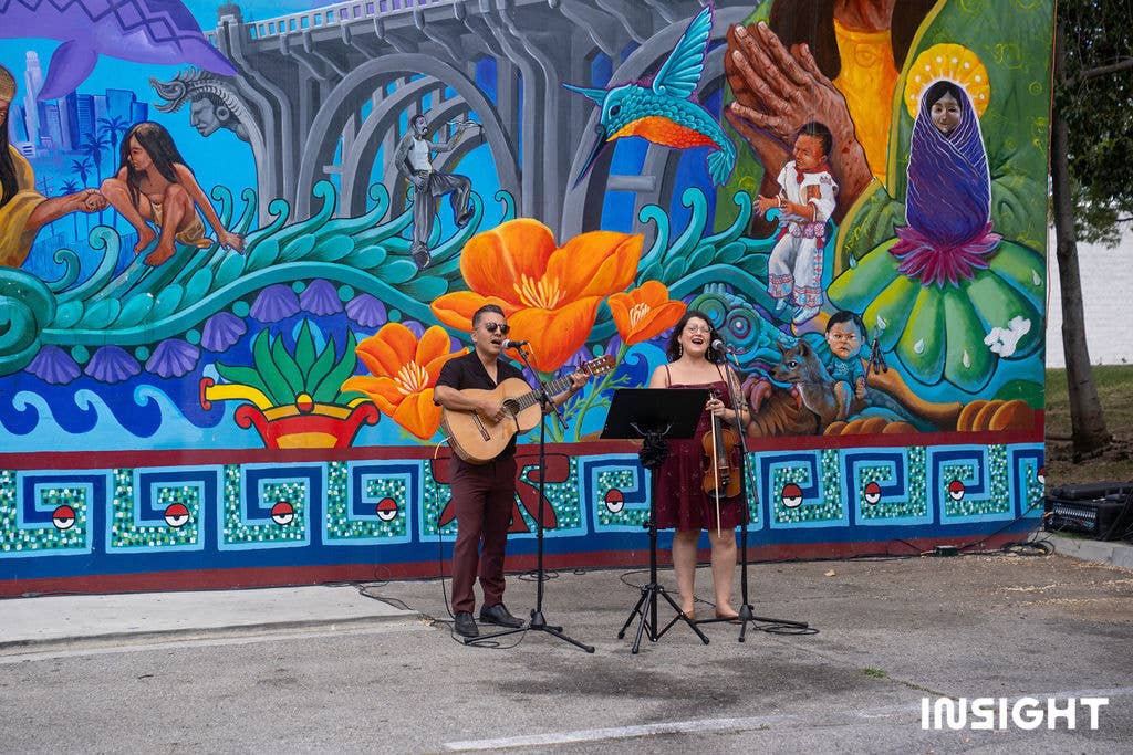 A guitarist and a violinist singing in front of a Great Wall of Los Angeles mural.