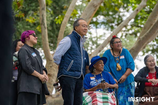 Yo-Yo Ma in blue outfit with Angelica Negron and the Tongva elders watching with a crowd of audiences outdoors in Riverfront Park.