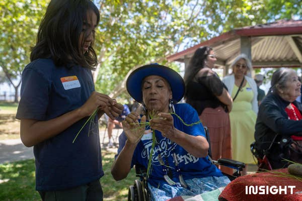 Woman in wheelchair shows younger boy how to braid using green yucca fibers.