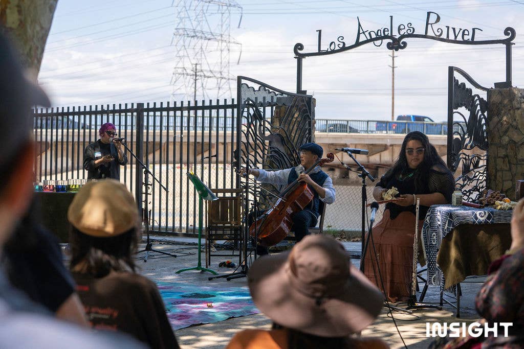 Yo-Yo Ma performing with two other people behind microphones in front of a gate to the Los Angeles River.