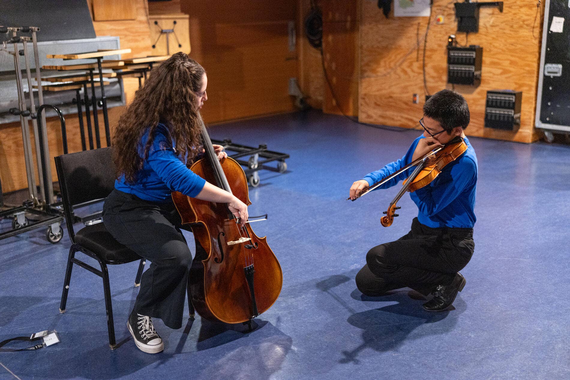 A cellist and violinist in blue YOLA National Festival shirts tuning in a green room.