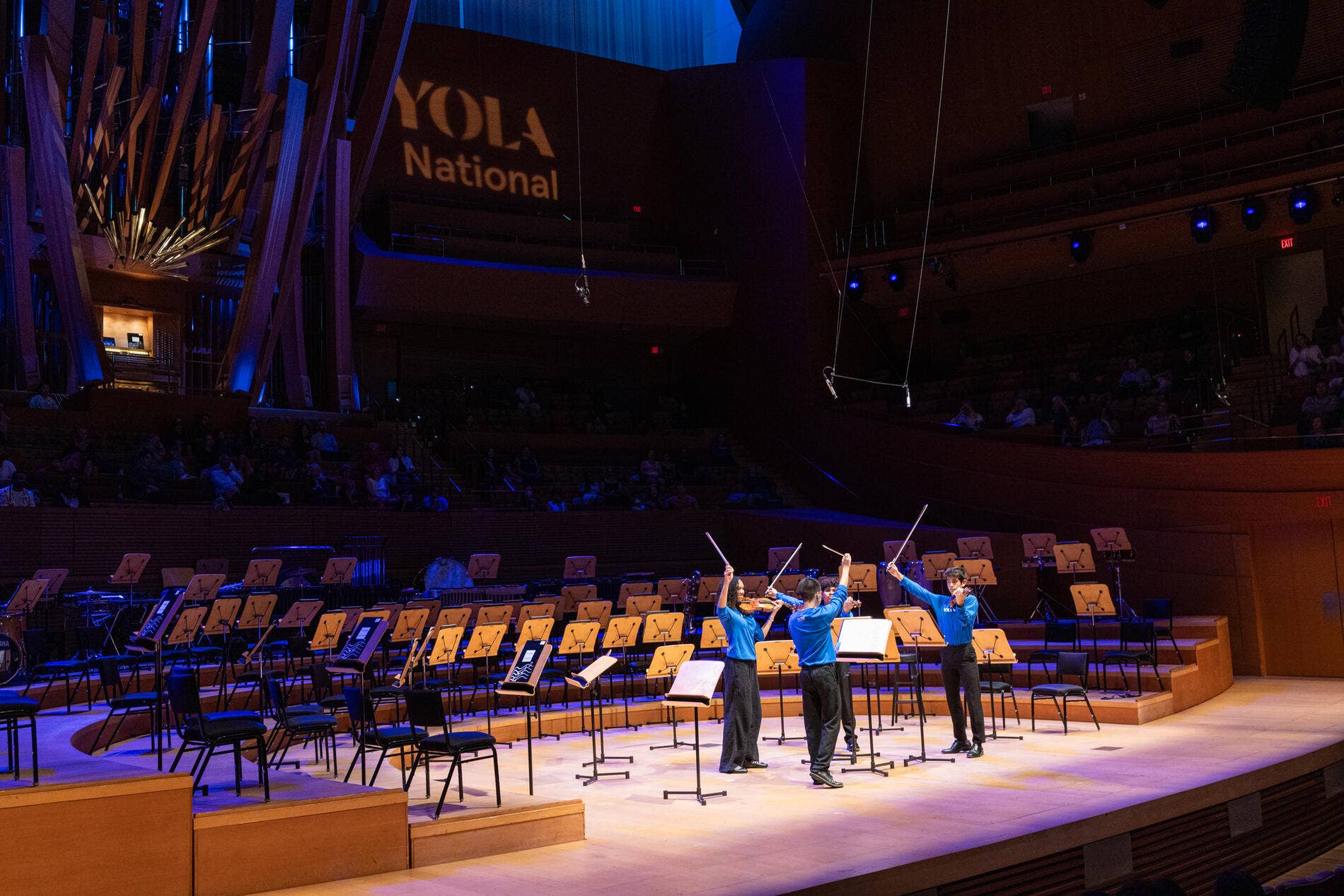 A string quartet dressed in blue YNF shirts lifting bows in the air on the Walt Disney Concert Hall Stage