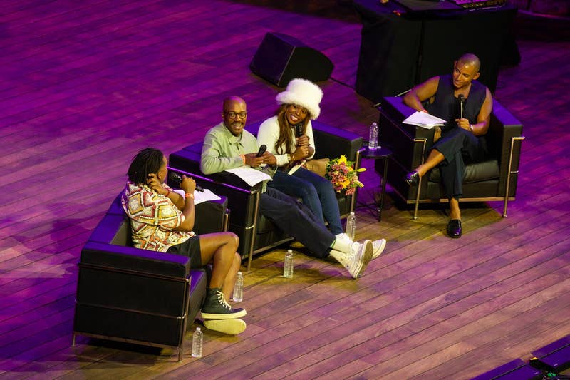 Panelists seated on stage with microphones and notes under purple lighting during a conference discussion.